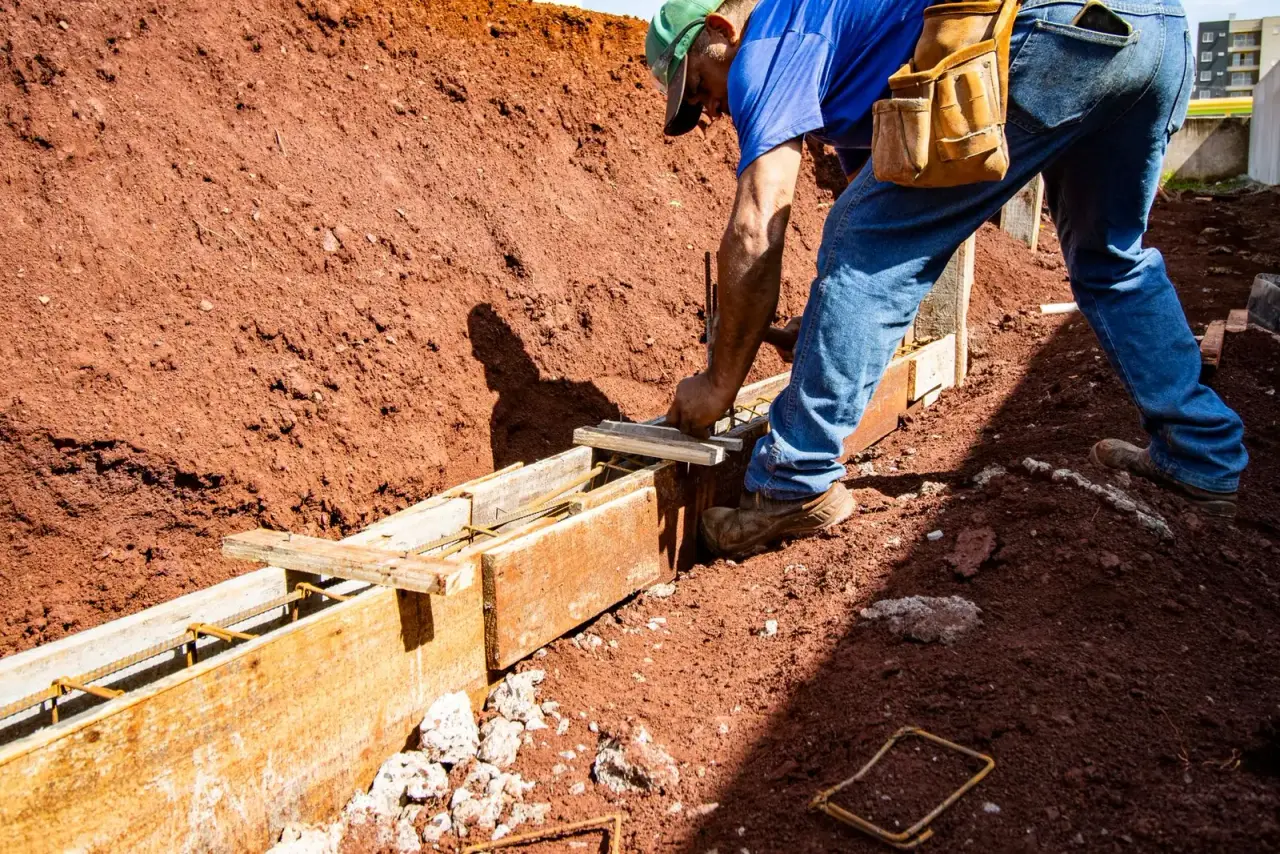 A worker digging a trench in red soil along a new concrete foundation