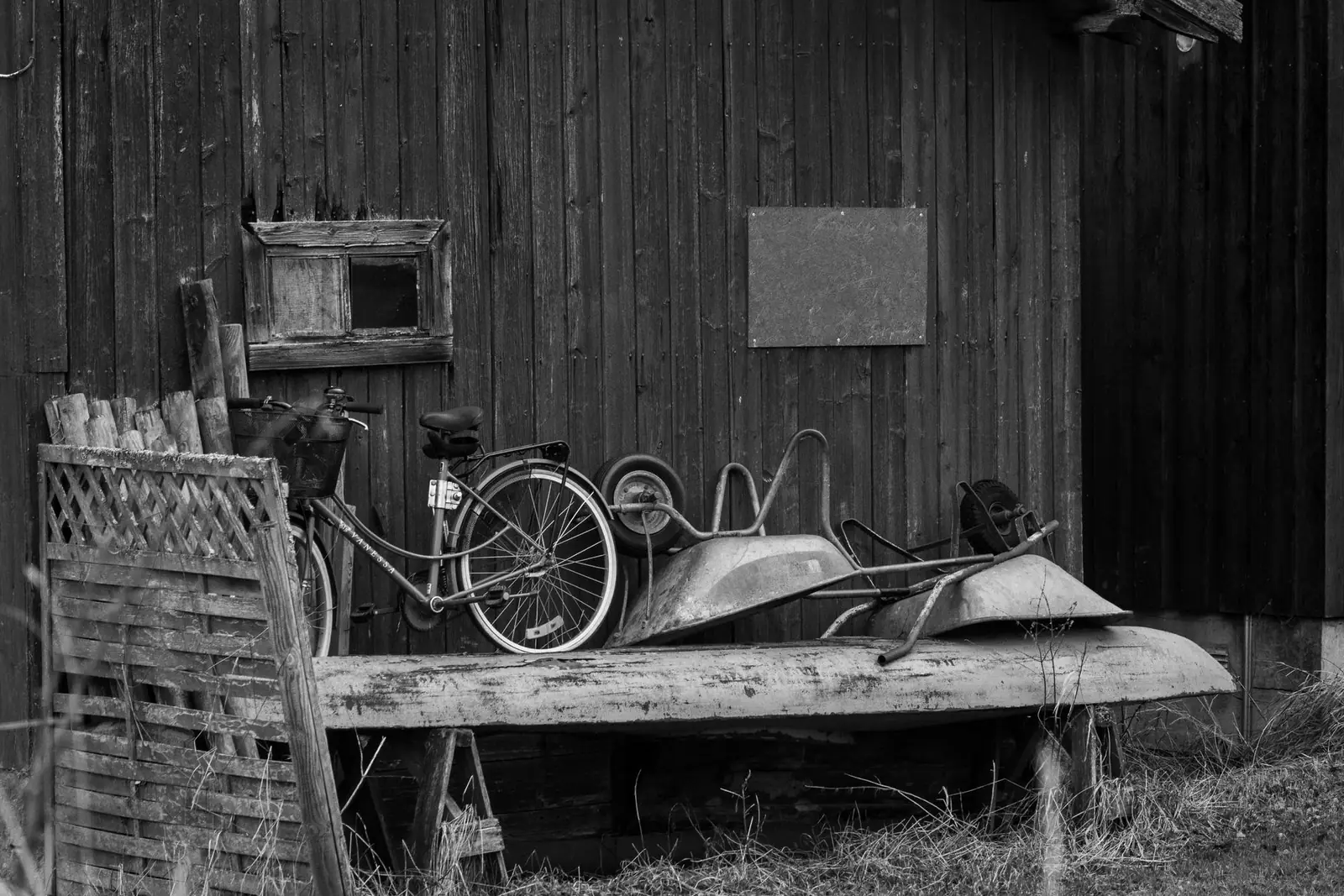 An old wooden shed with a bicycle and a rusty wheelbarrow outside, in black and white