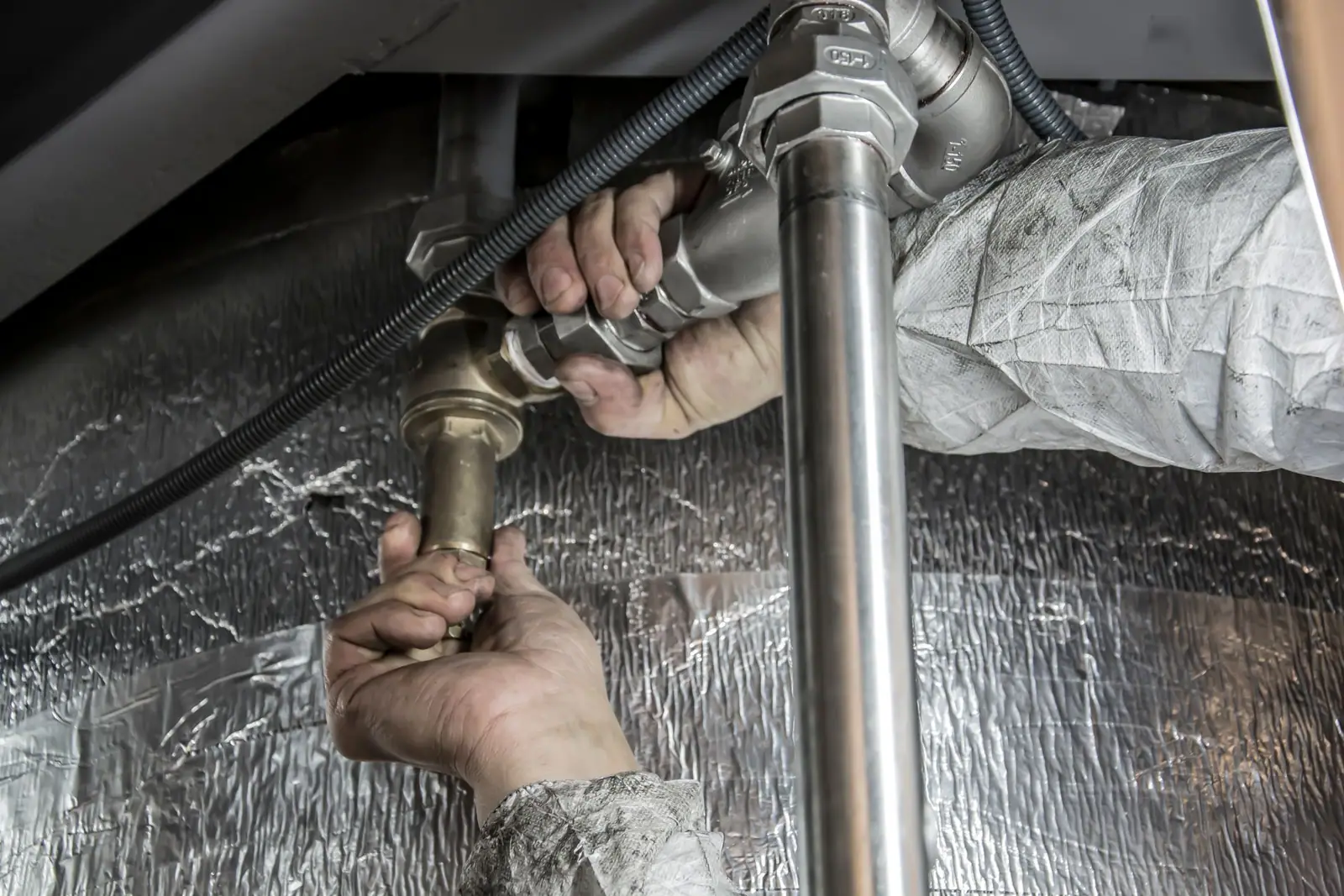 A plumber's hands adjusting metal pipe fittings beside an insulated wall