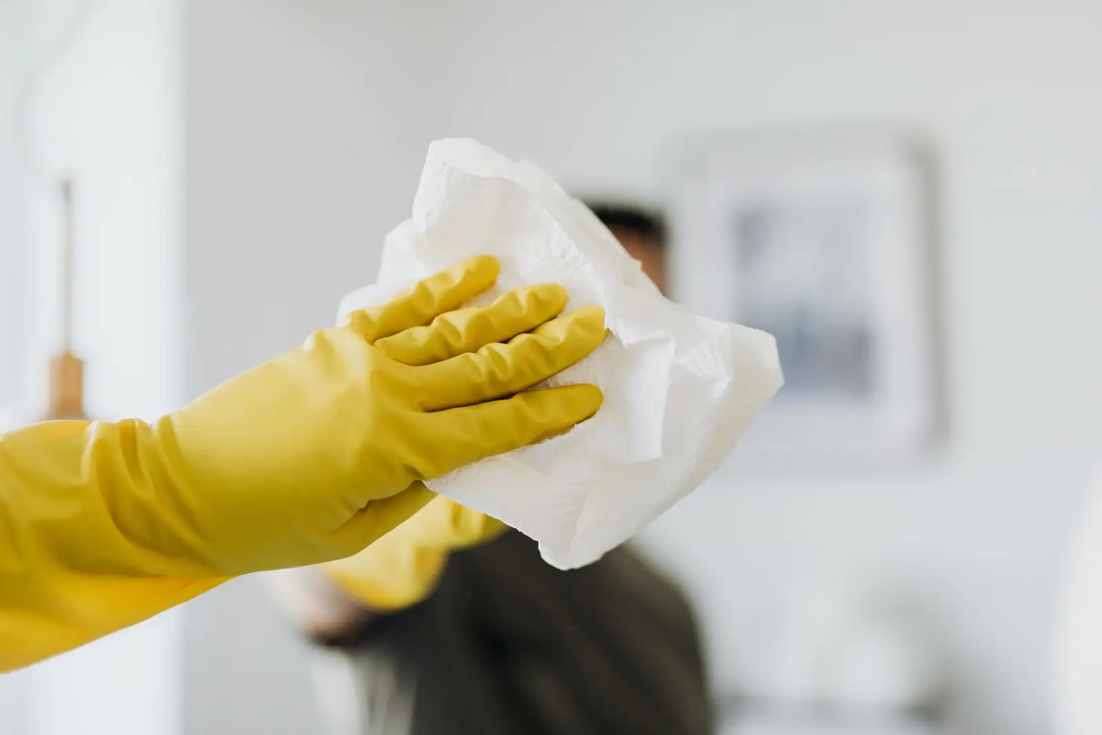 A person in yellow rubber gloves wiping a bathroom mirror with a paper towel