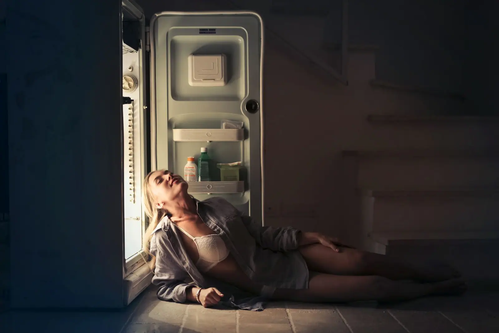 A person sitting on the floor beside an open refrigerator in a dark room
