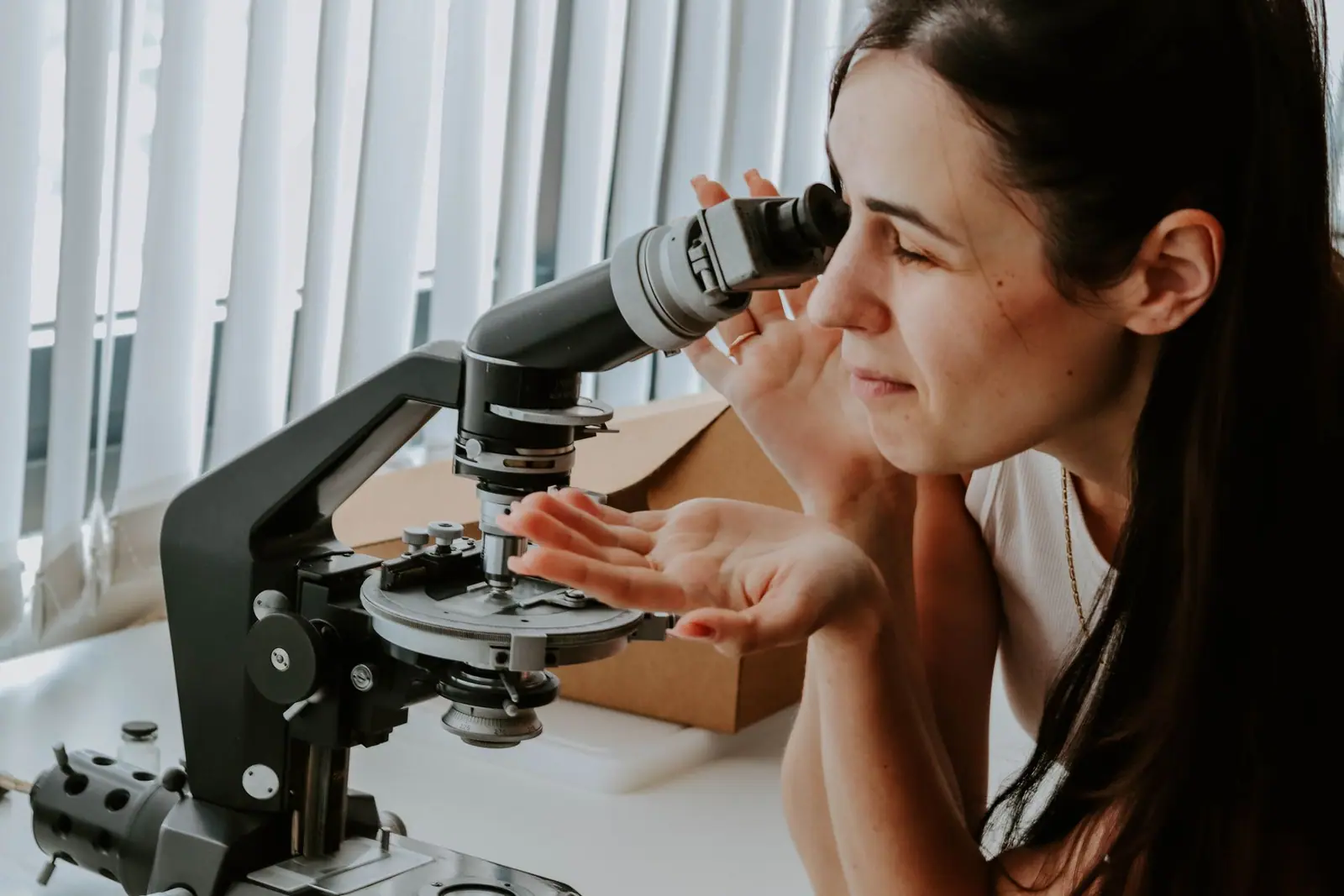 A woman looking through a laboratory microscope with one hand near its controls