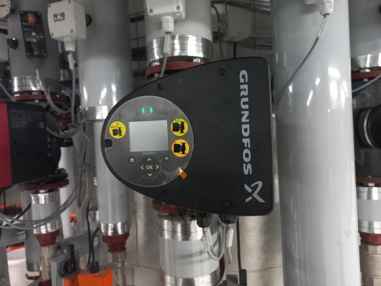 A black Grundfos pump control mounted among pipes in a swimming pool equipment room