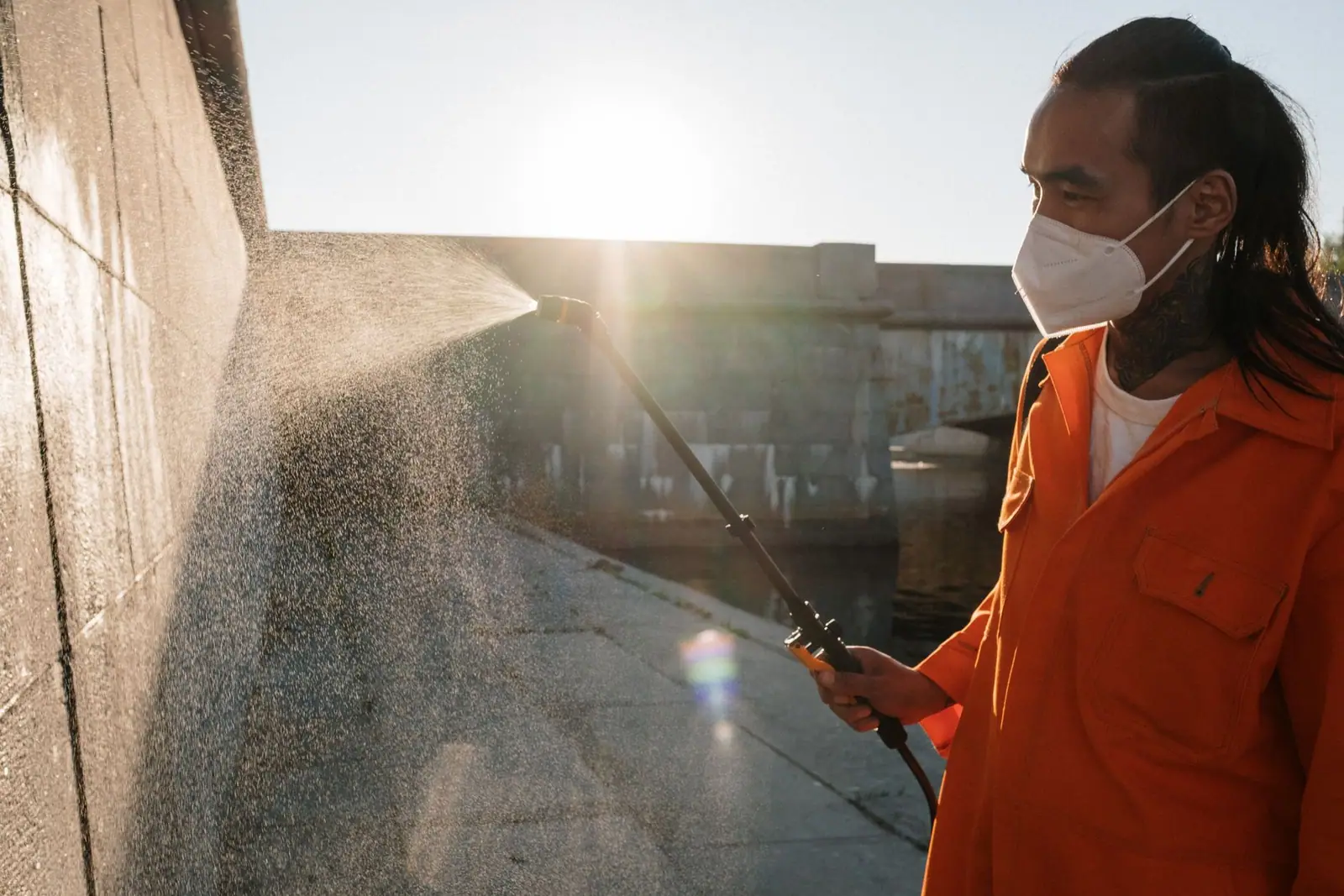 A person in an orange uniform and mask spraying a wall with a cleaning lance