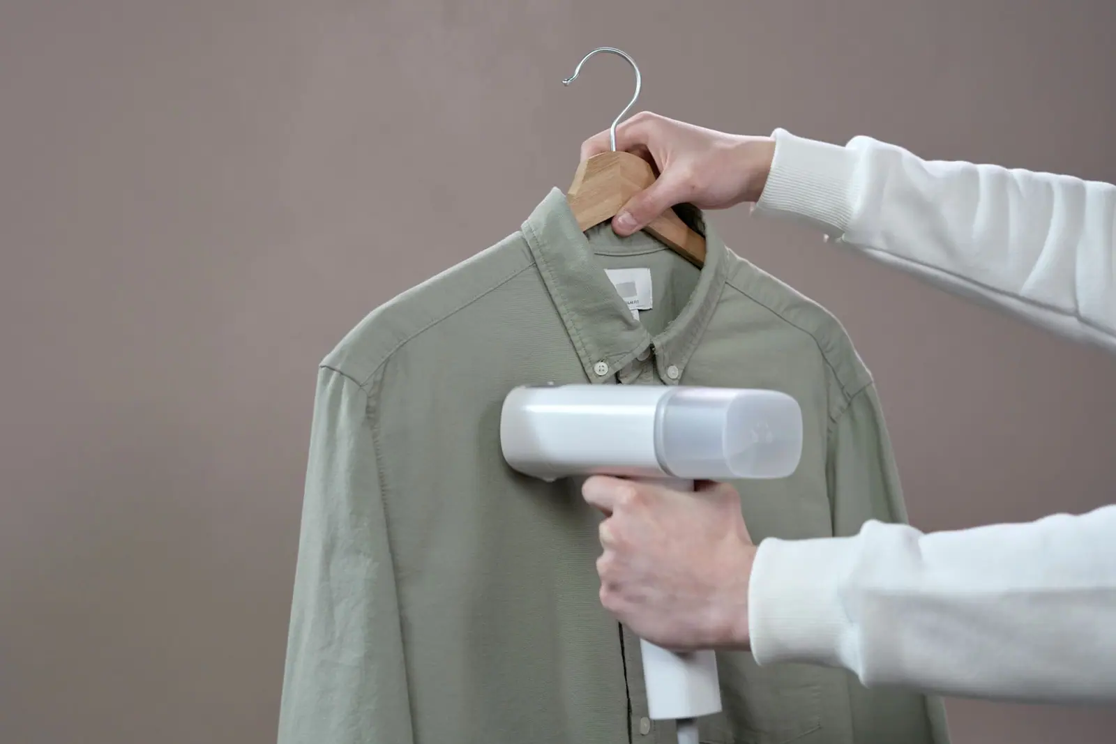 Hands running a handheld garment steamer over a green shirt on a hanger